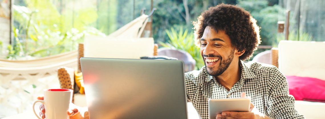 homem negro, sorrindo, sentado em um ambiente ensolarado de uma casa, trabalhando em home office